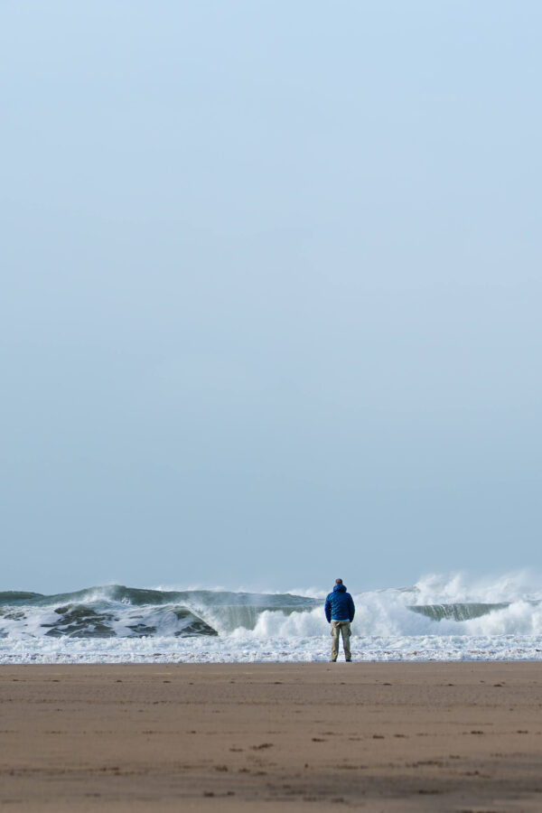Man in a blue jacket standing alone on a beach watching large waves breaking on the shore